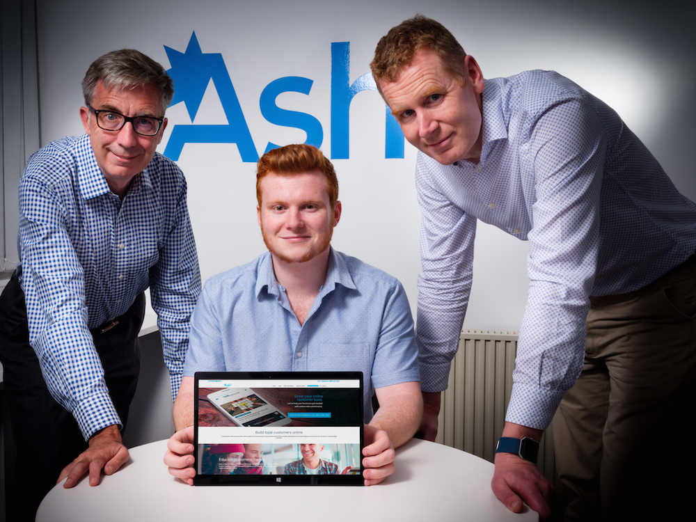 3 men from Ash TV gathered around a table showing an Ipad with their website displayed