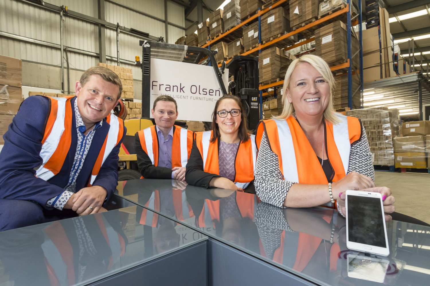 Four women standing in a factory with Frank Olsen signage behind them. They are all wearing high vis jackets. There are two women in the group and the woman to the far right has blonde hair and standing next to her is a woman with dark hair wearing glasses.
