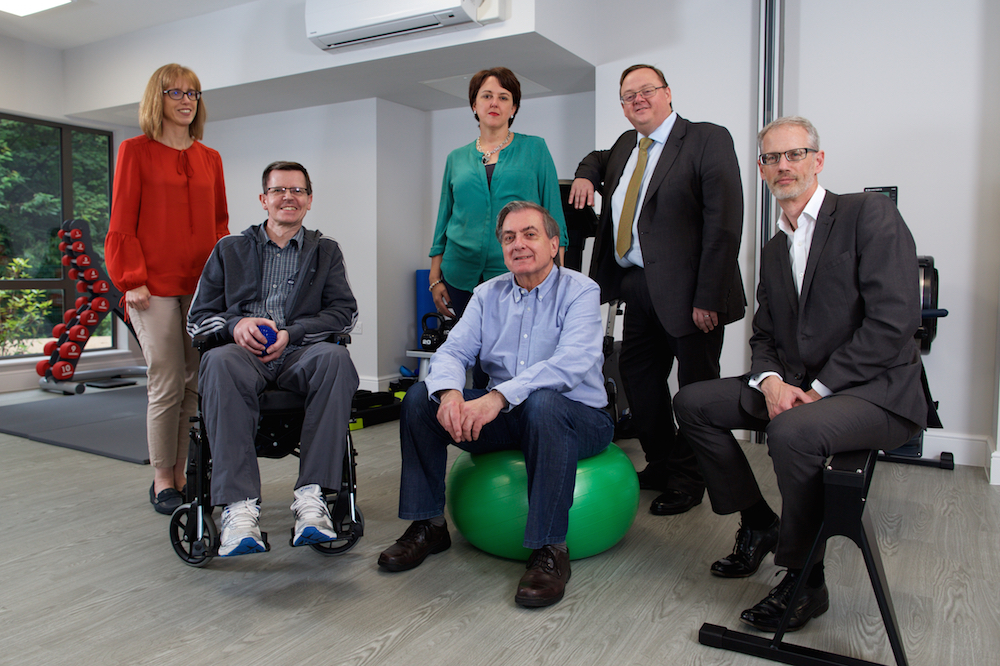 Two women wearing casual clothes, one man in a wheelchair, one man sitting on a large exercise ball and two men wearing black suits.