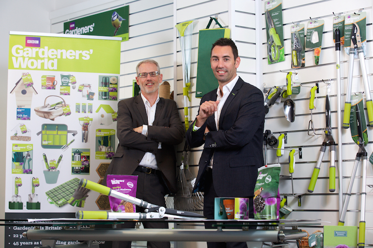 Two suited men surrounded by gardening equipment with a large Gardeners World point of sale poster behind them.
