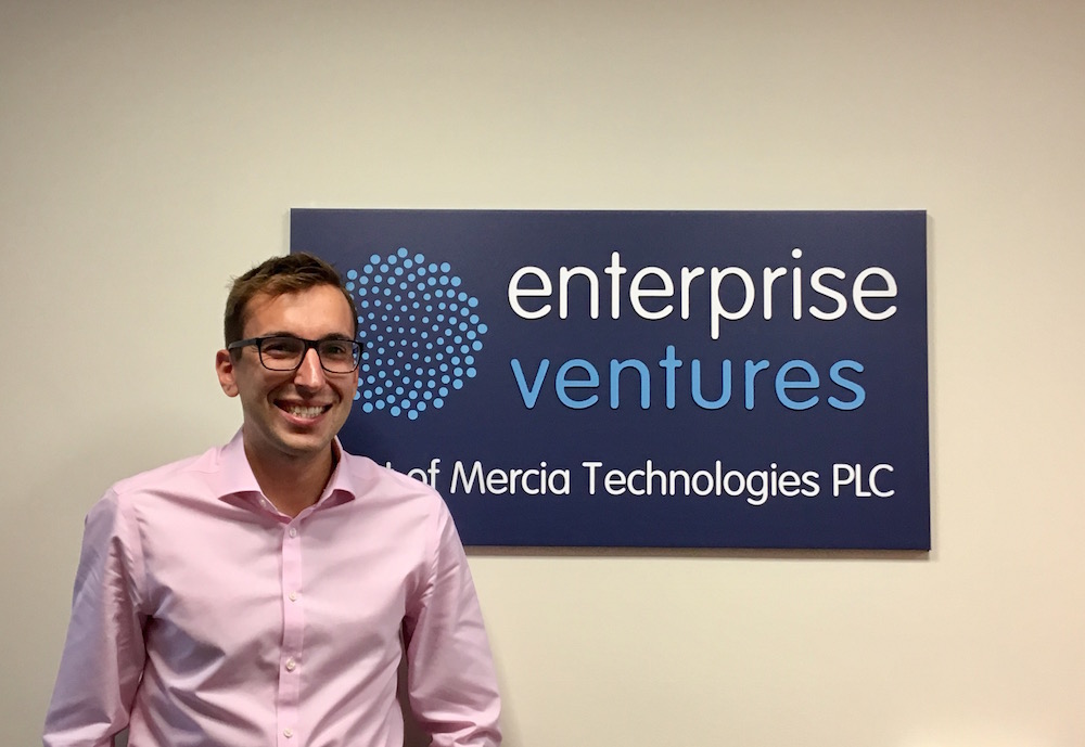 Man with glasses wearing a pink shirt standing in front of wall and signage stating Enterprise Ventures.