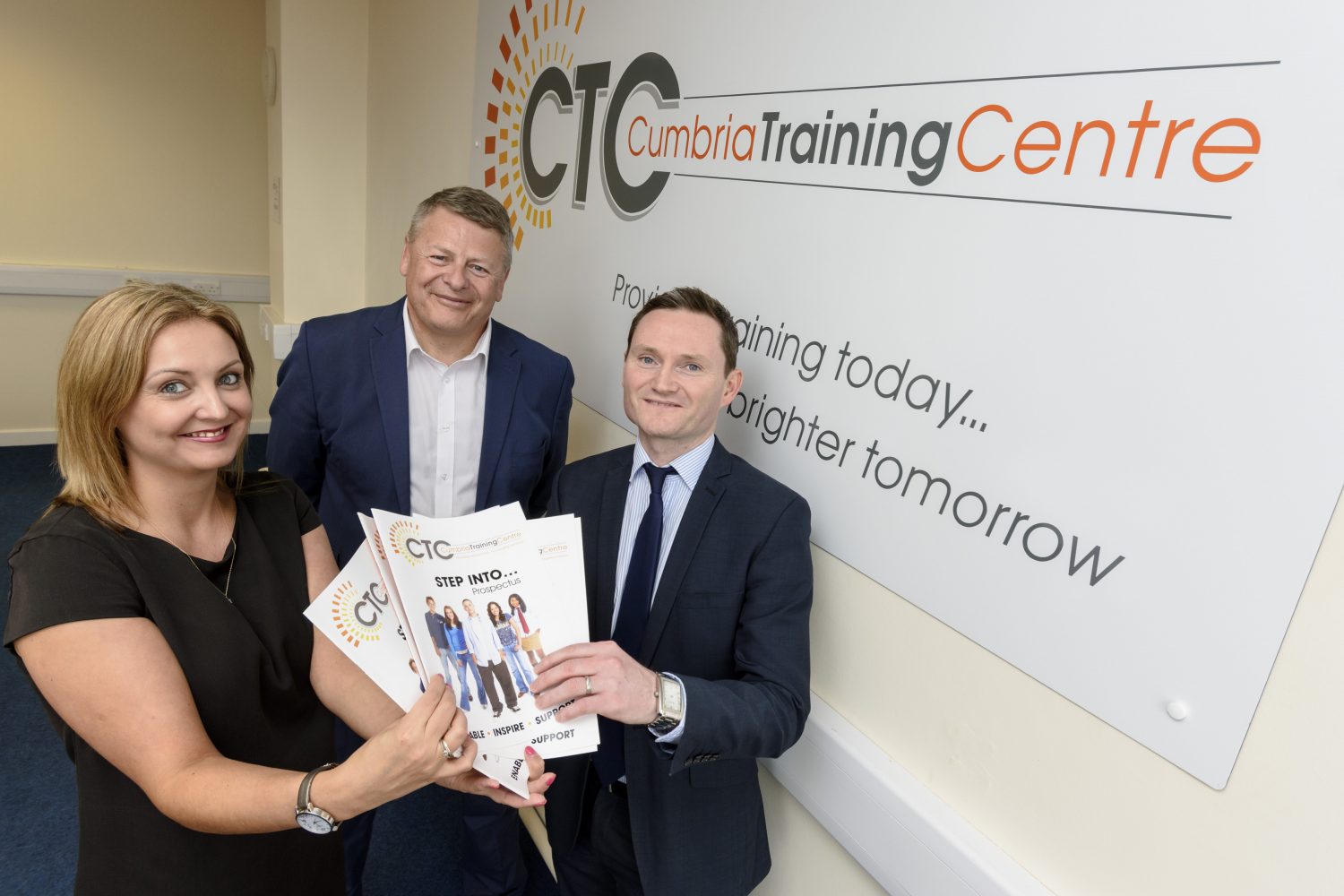 Three people in an office standing near a wall with a poster of CTC Cumbria Training Centre poster. There is one woman to the far let with light brown hair wearing a black tee shirt holding some brochures and another man to the far right wearing a blue suit and blue tie also holding the brochures.