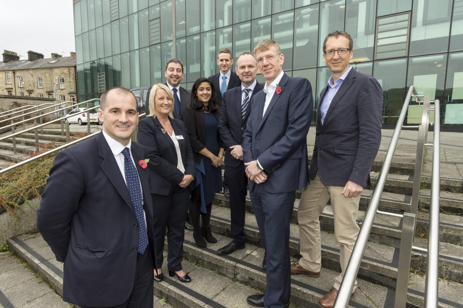 Eight people standing on externa steps. Man standing by himself is wearing a blue suit with a Rembrance poppy on it has a blue patterned tie on, there are two women in the group. One woman has blonde hair and is wearing a suit and the other woman has dark mid length hair.