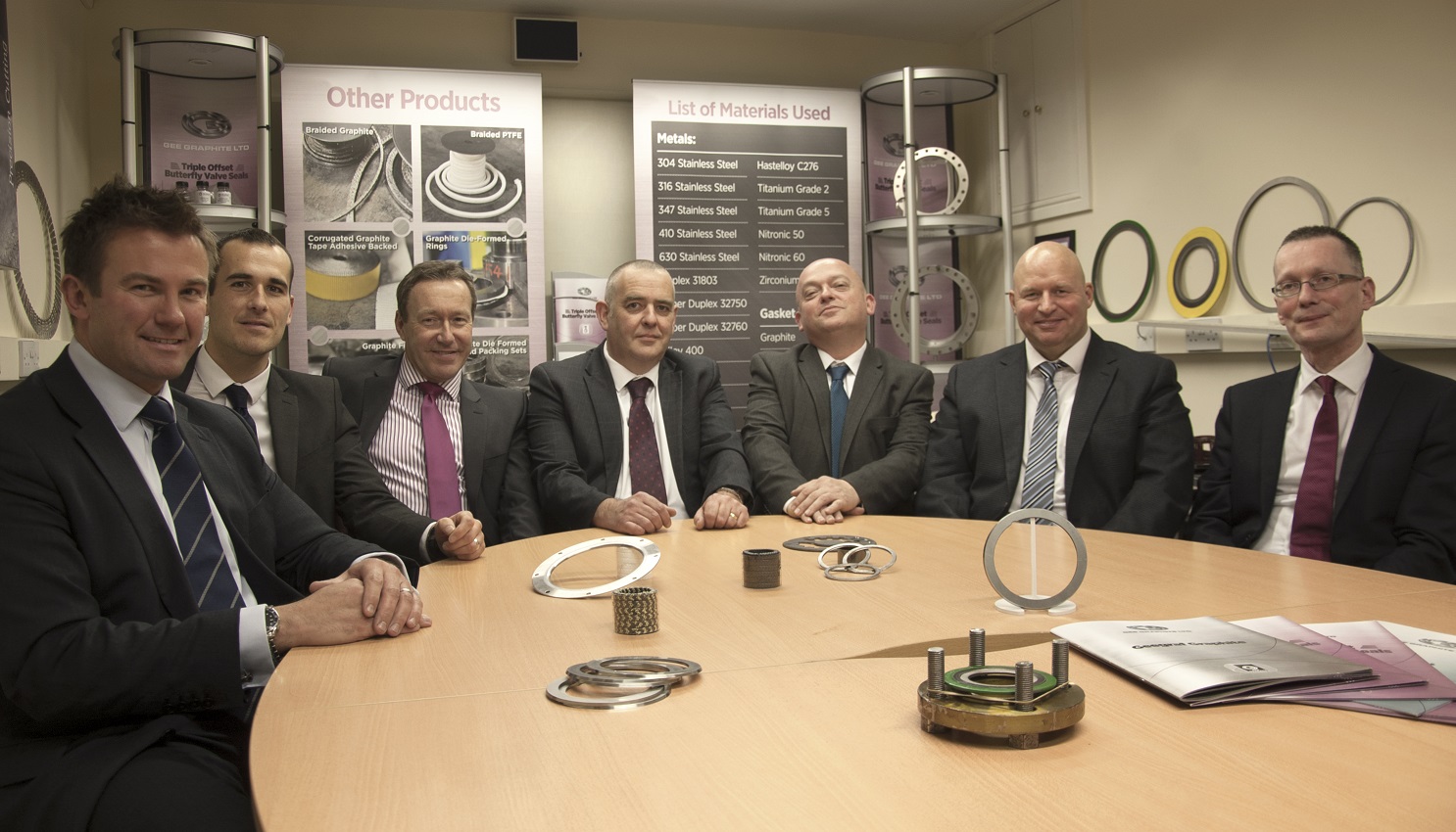 Seven suited men sitting around an oval table surrounded by industrial products on the table and point of sale behind them.