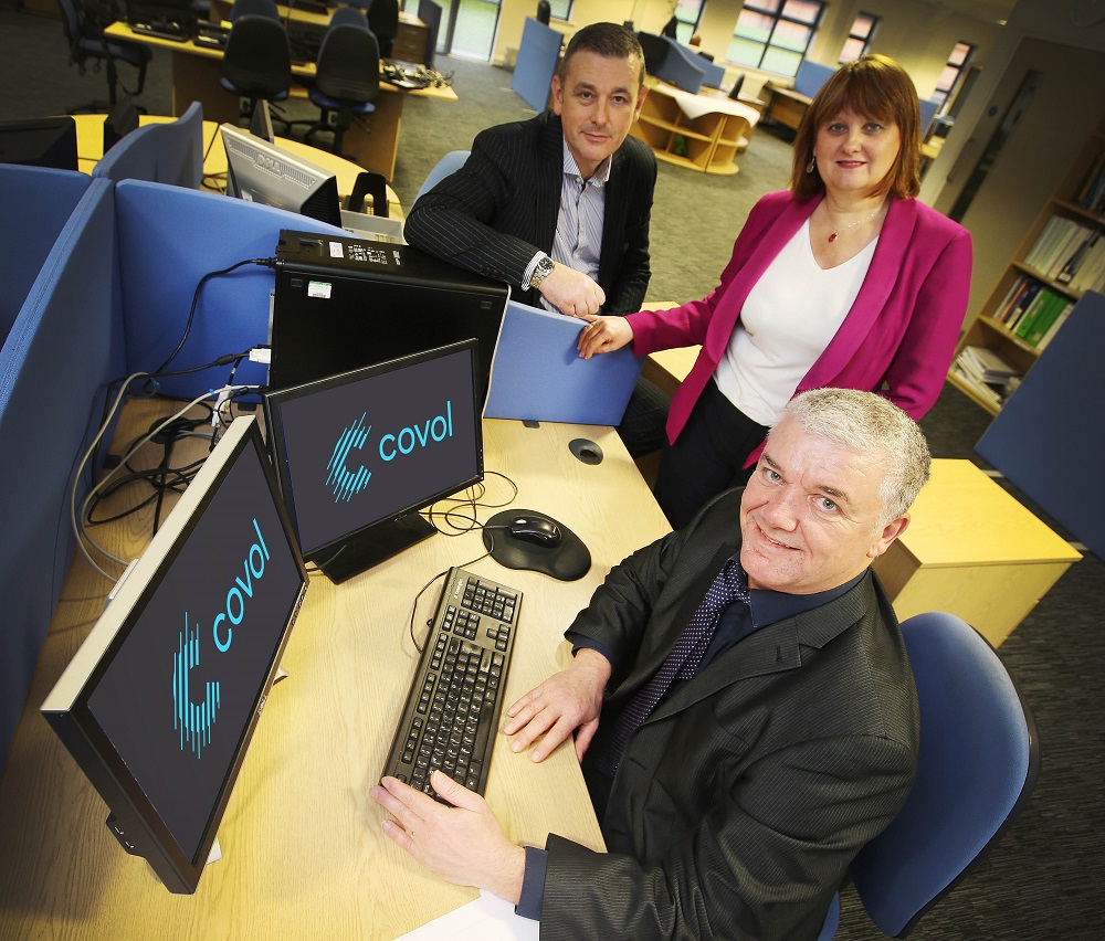 One business man sitting at a desk in front of two computer screens with Covol branding on it. There is a woman and man standing around the desk.
