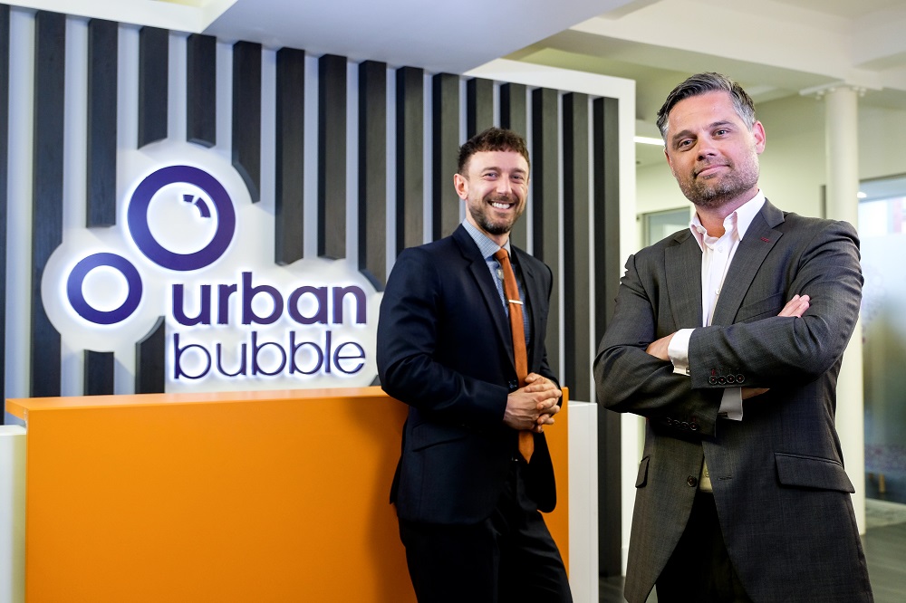 Two business suited men standing in front of an orange painted desk with Urban bubble branding on the wall behind them.