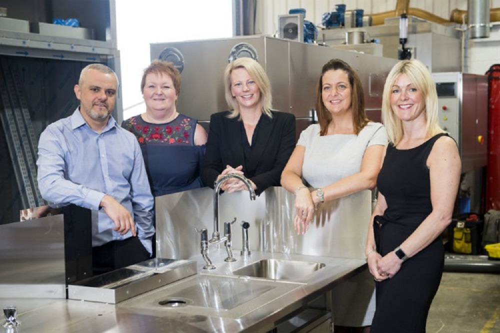 Group of four women in factory and one grey haired bearded man in the far left. They are standing around an industrial sink.