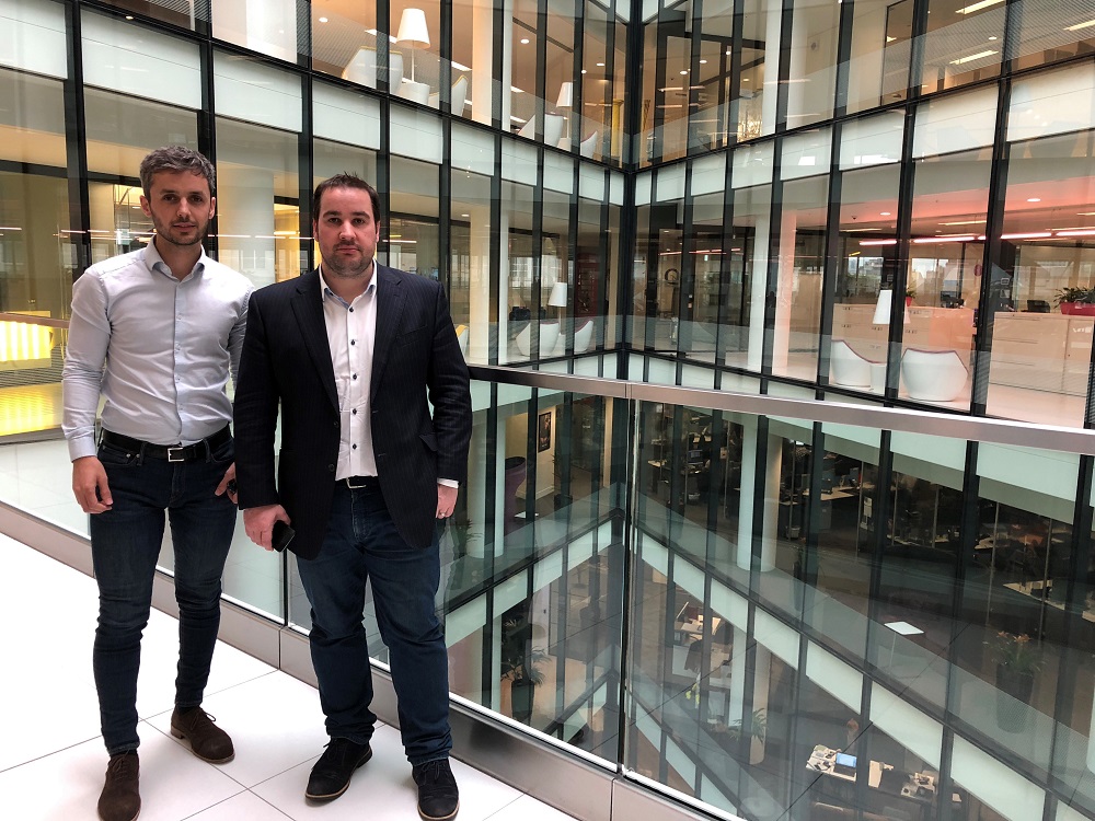 Two businessmen standing in an office building. They are in front of a large glass wall.