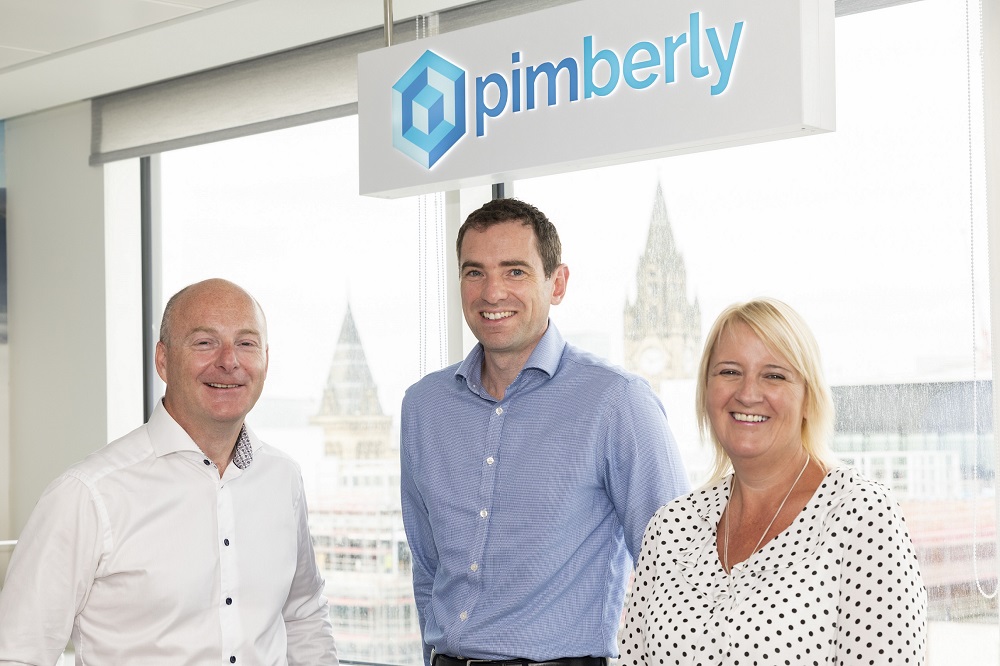 Three people in an office standing in front of a window with signage stating Pimberly. The woman to the far right is wearing a white and black spotted top and has blond hair. The man in the middle is wearing a blue shirt and the man to the far left is wearing a white shirt.