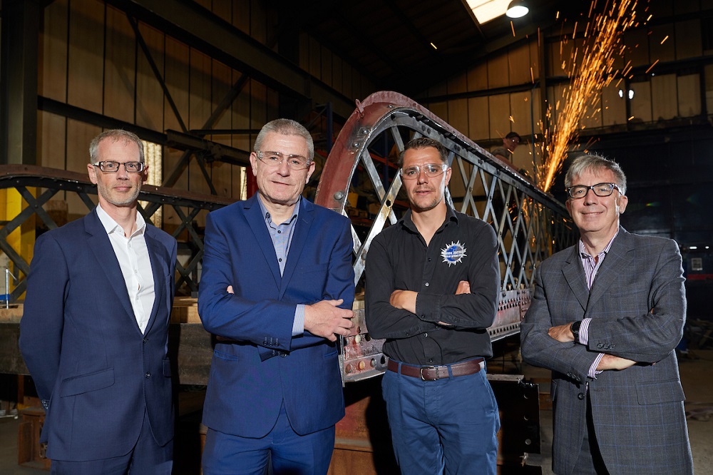 Two men in blue suits , one man in blue jeans and a navy blue shirt and man to far light in a grey suit in an industrial setting.