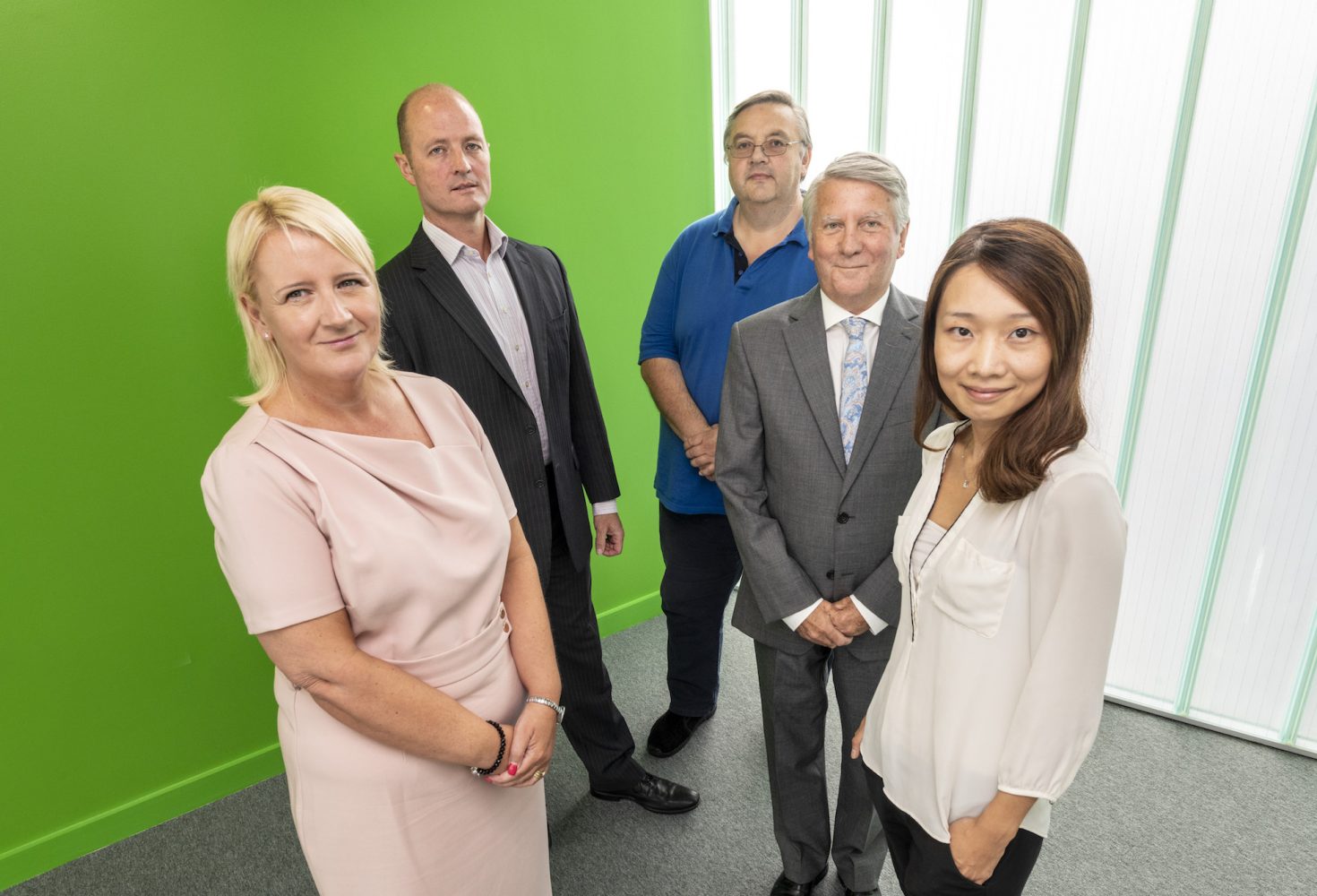 Five people in an office standing in front of a bright green wall. There are two women and the woman to the far left has a peach coloured dress on and has blonde hair. The additional woman on the far right is Asian and has long dark hair and is wearing a white shirt.
