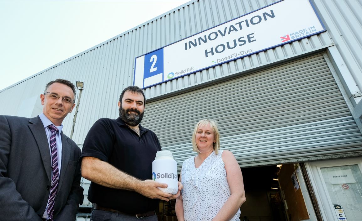 Two men and a woman with blonde hair standing outside office named Innovation House. Man in the middle of the group holding a plastic canister with Solid Tek on it.
