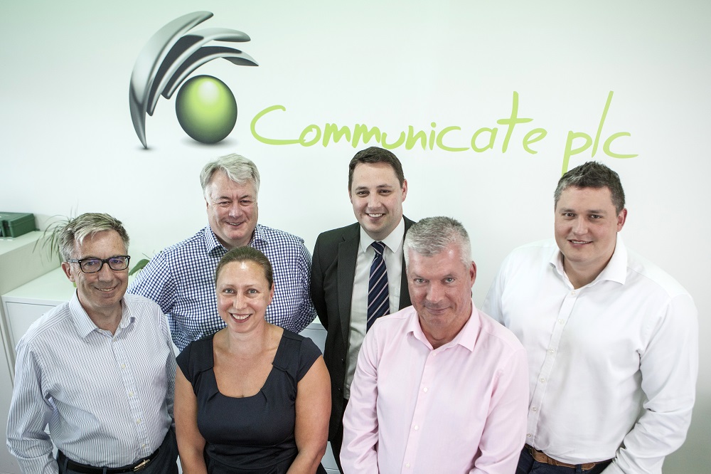 Employees from Communicate Technology smiling and stood in front of a Communicate plc branded wall