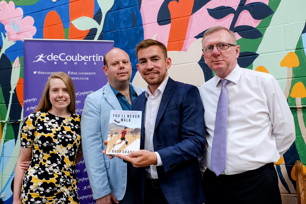 Three men and one woman in a colourful dress standing in front of a colourful brick wall. Man in the middle of the row of people is holding a book called 'You'll New Walk'.