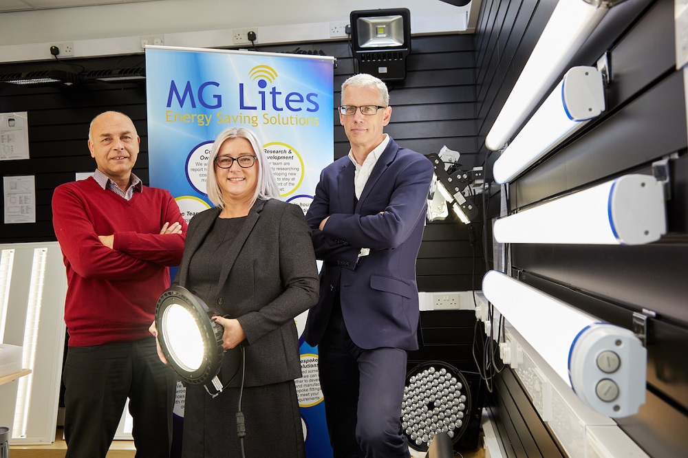 One man wearing a red jumper, woman wearing a brown suit and man with navy blue suit standing in front of MG Lites signage.