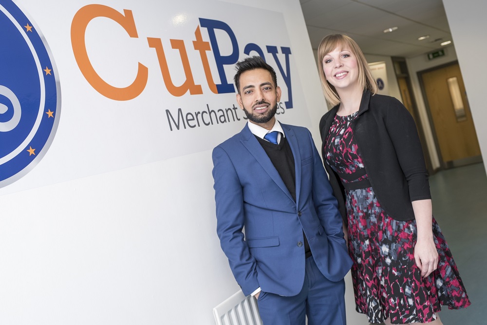 A man and a woman stood in a office building in front of a CutPay Merchant Services branded wall