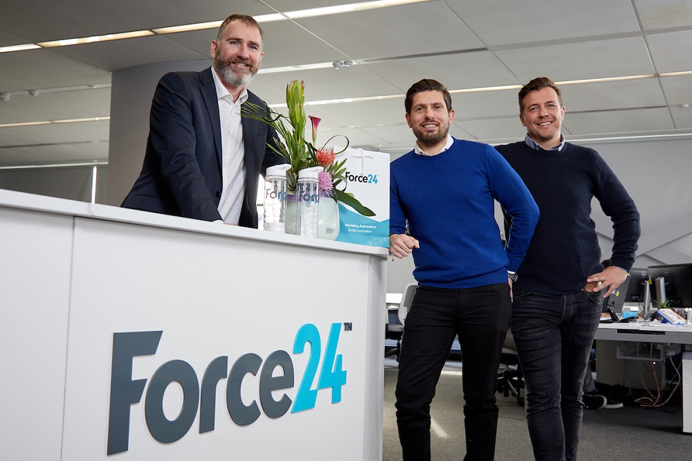 Three men in an office. One man standing behind a workstation with Force24 written on it.