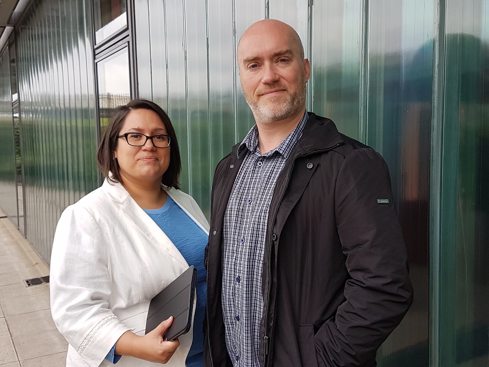 Man and woman outside green industrial building. Woman wearing glasses and has dark shoulder length hair to the left of the man is wearing a white jacket and a light blue top.