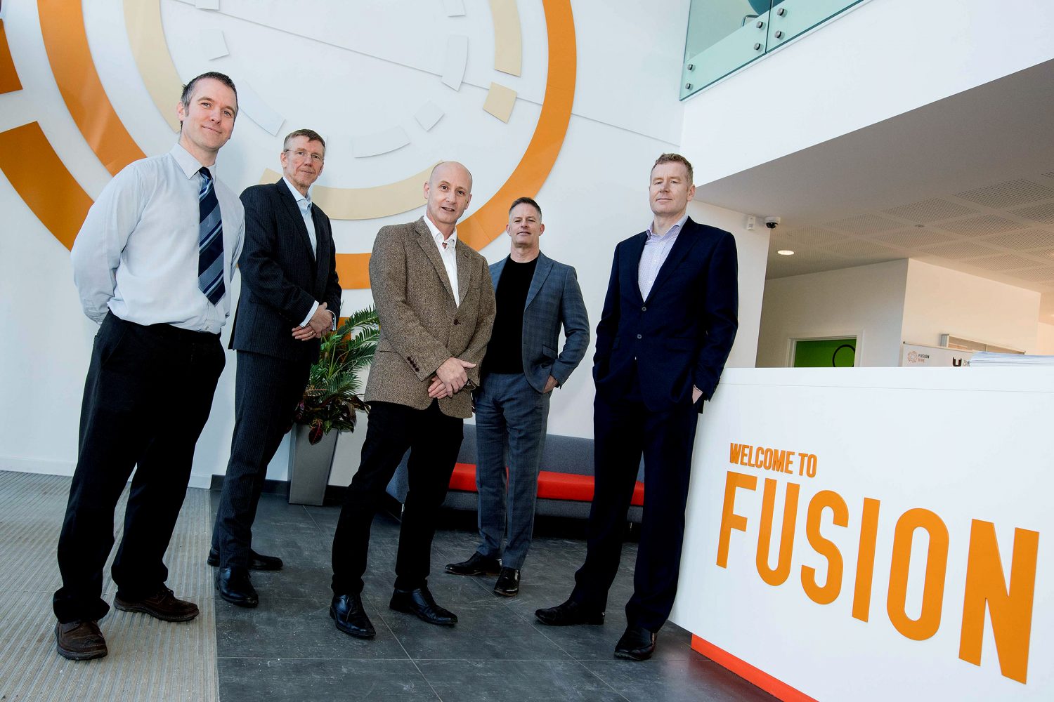Five men standing, man to far right wearing a black suit is standing against a reception desk with Welcome to Fusion signage on it.