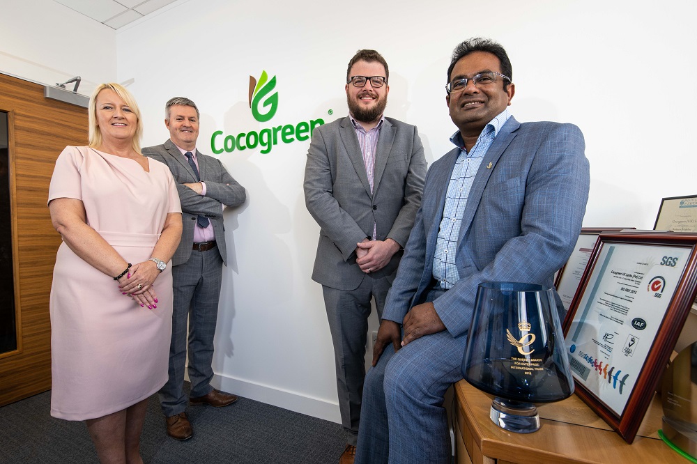 Group of four people in a office room standing in front of a wall with the Cocogreen logo. There is only one woman and she has blonde hair and is wearing a pink dress. There is an Asian man to the far right wearing a blue suit and a blue checked shirt. He is sitting on a desk surrounded by awards.