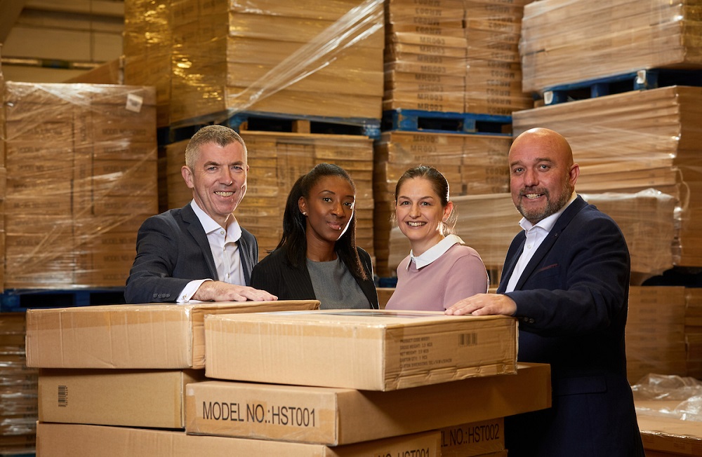 Two men and two women in a factory standing next to lots of cardboard boxes. Man to the far right is bald and has a beard, he is wearing a navy blue suit and white shirt, he is standing next to a lady wearing a dusty pink jumper, the next lady has dark skin and is wearing a black jacket and grey top and finally to far left there is a man wearing a black suit and white shirt.