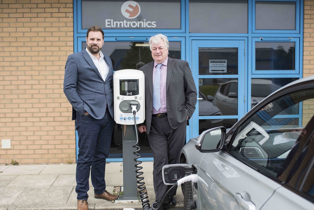 Two men standing behind building with Elmtronics signage. Man to the left is standing next to an electric charging post for vehicles is wearing a blue suit and white open necked shirt. He has a beard and dark hair. The man to the right has grey hair and is wearing a grey suit.