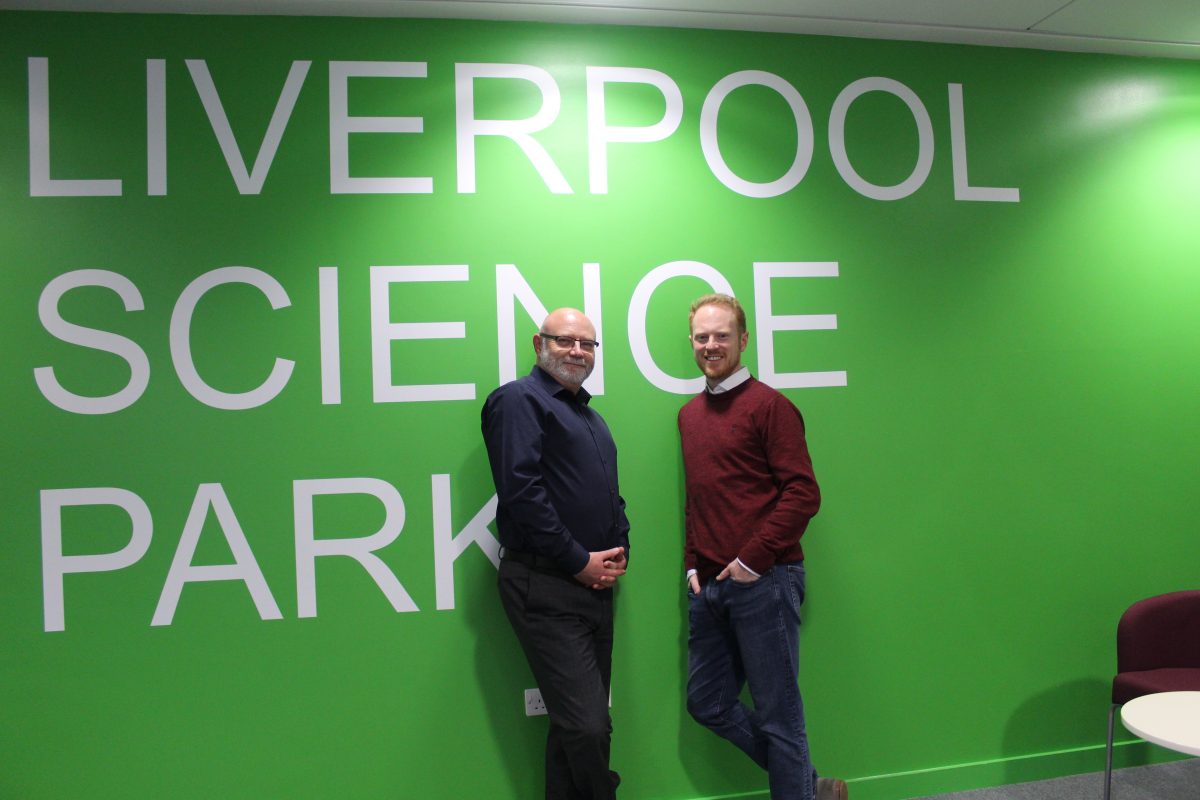 Two men standing in front of a green wall with white writing stating Liverpool Science Park. Man to the left has glasses andis wearing a dark blue suit and man to the right is wearing a maroon jumper and blue trousers.