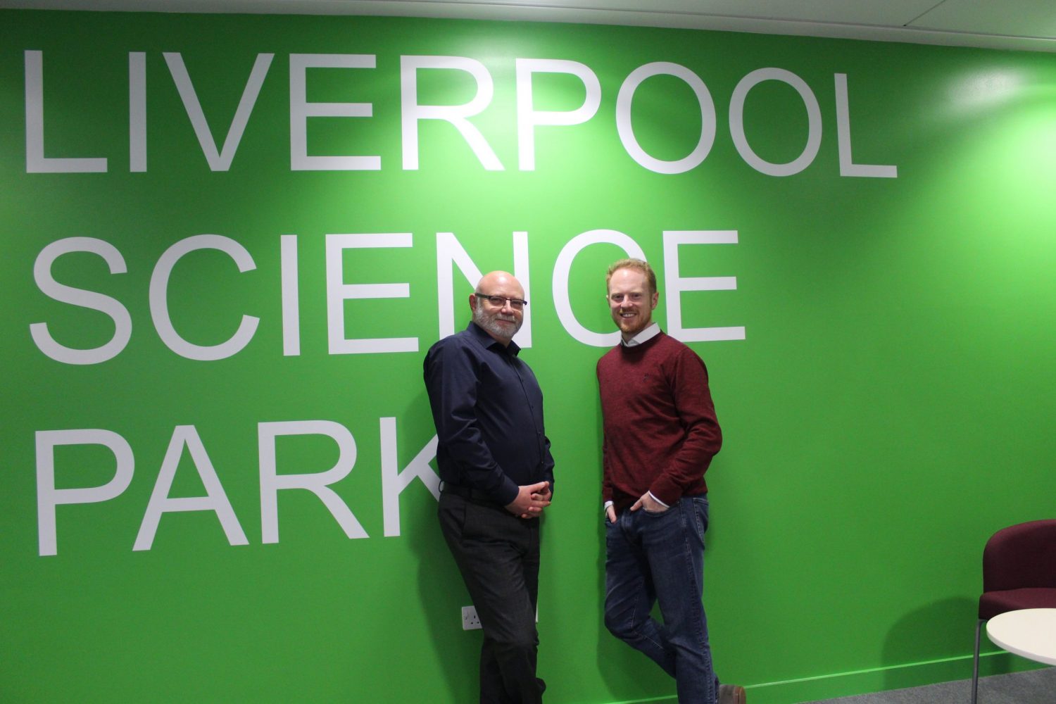 Two men standing in front of a green wall with white writing stating Liverpool Science Park. Man to the left has glasses andis wearing a dark blue suit and man to the right is wearing a maroon jumper and blue trousers.
