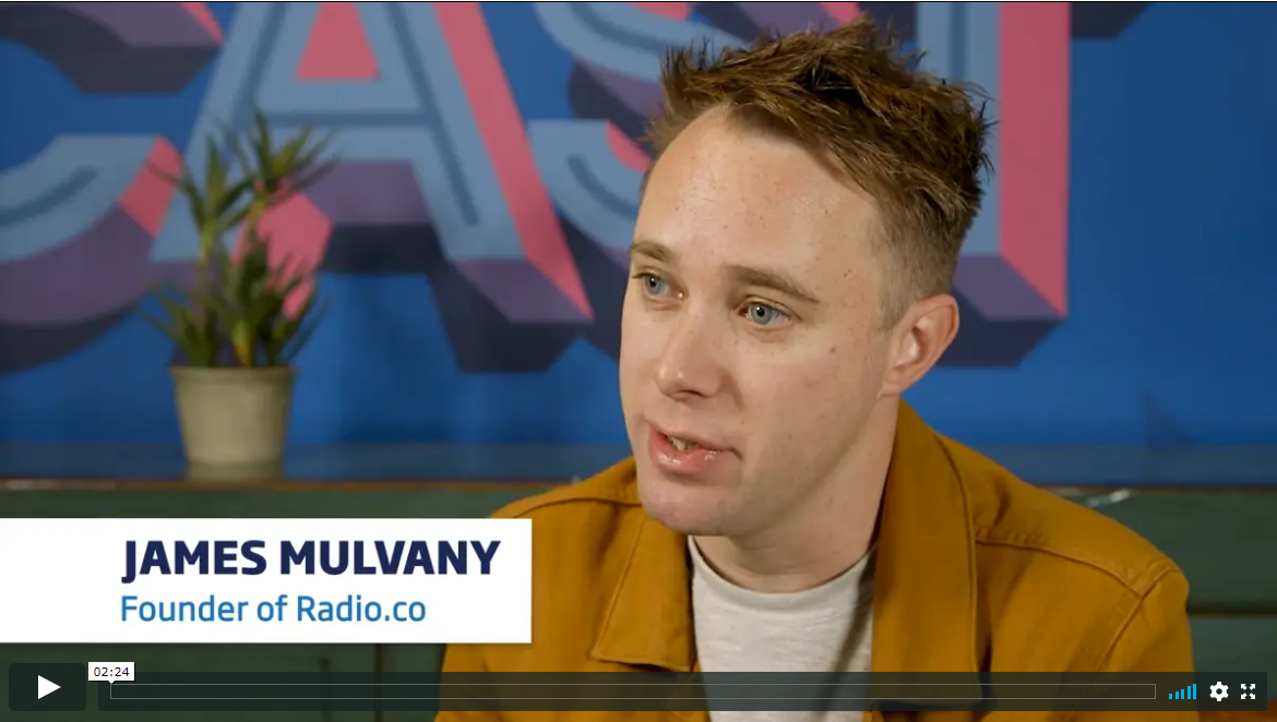 Young man sitting behind a colourful wall is wearing a mustard yellow jacket and a beige sweat shirt. He has blue eyes. He is named James Mulvany and is the founder of Radio.co