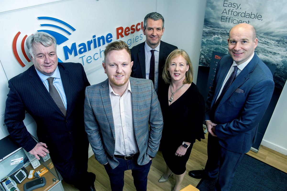 Five people standing in front of Marine Rescue Technologies poster in an office. Man to the far left is bald and is wearing a blue shirt with a white shirt and dark navy tie, woman to the left of him is wearing a black dress. The remaining people are men. There is one man directly behind the woman wearing a black jacket and has a black tie and a man to the far left leaning against the Marine Rescue Technologies poster wearing a navy suit with a light blue shirt.