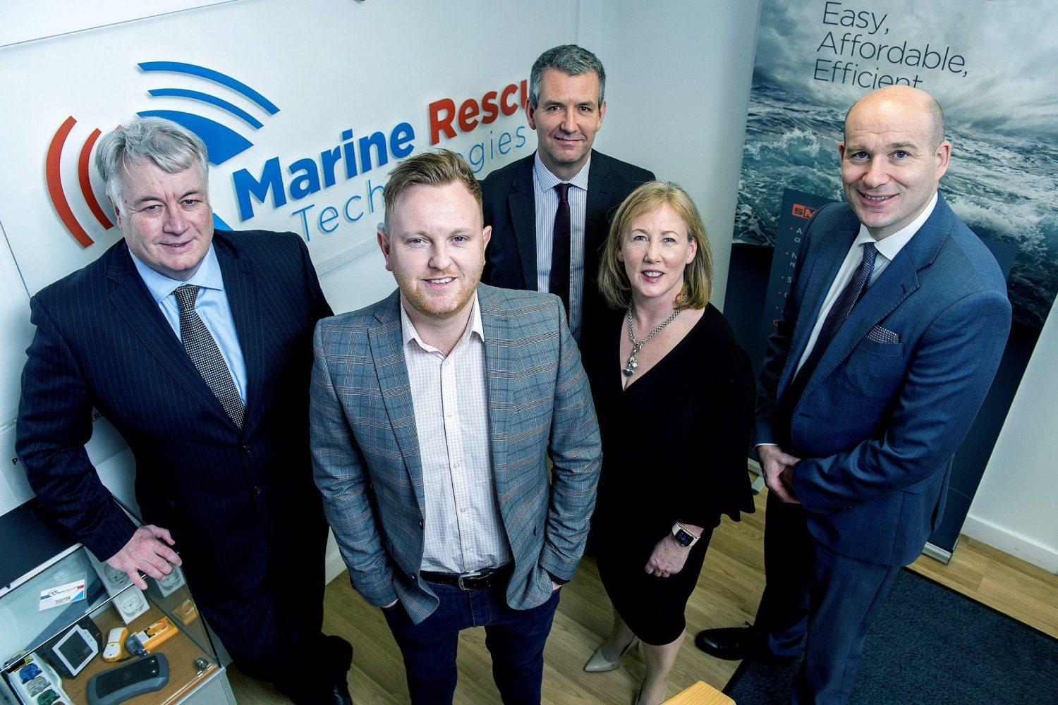 Five people standing in front of Marine Rescue Technologies poster in an office. Man to the far left is bald and is wearing a blue shirt with a white shirt and dark navy tie, woman to the left of him is wearing a black dress. The remaining people are men. There is one man directly behind the woman wearing a black jacket and has a black tie and a man to the far left leaning against the Marine Rescue Technologies poster wearing a navy suit with a light blue shirt.