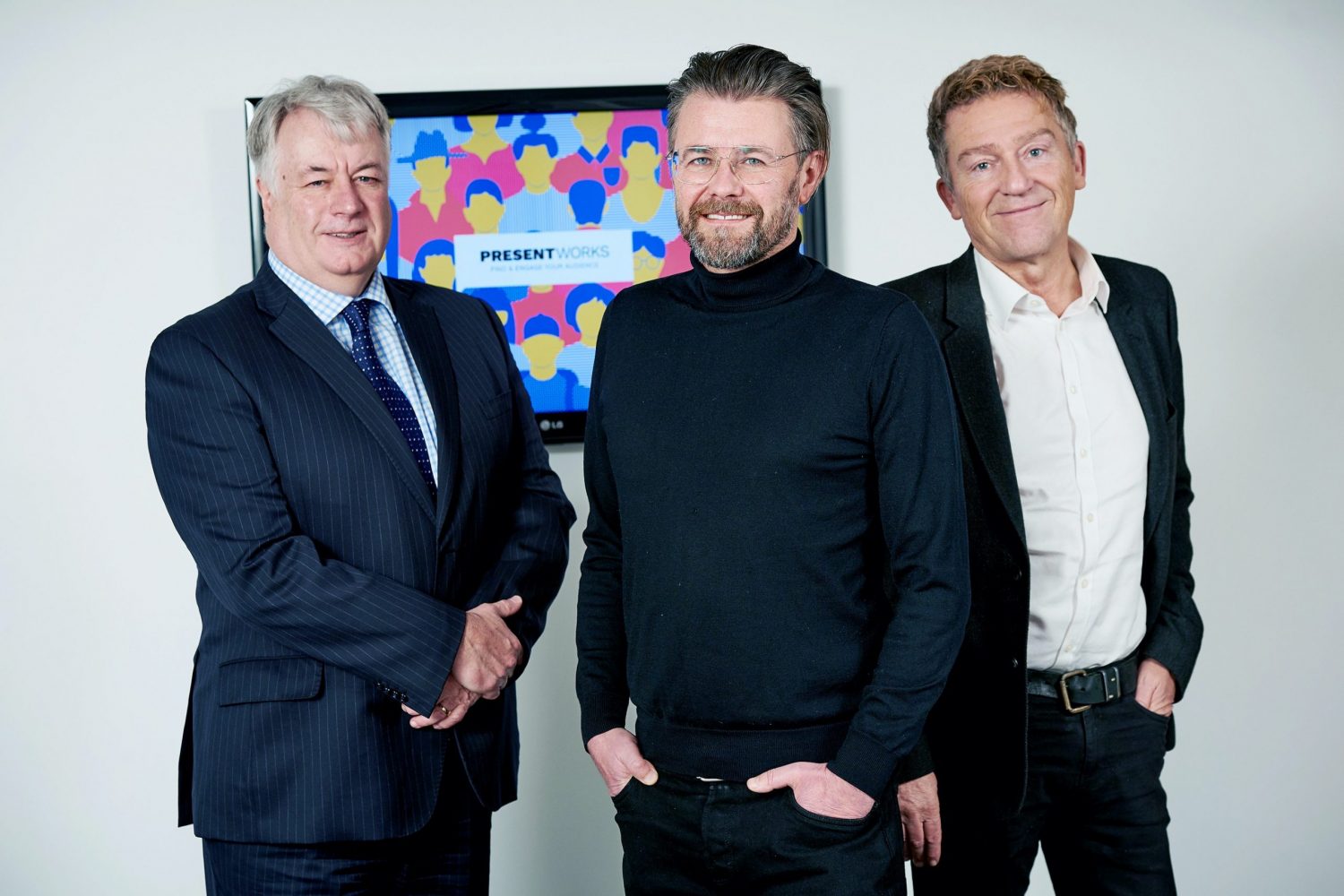 Three men standing in front of screen showing Present Works on the screen. Man to the far left is wearing a black suit with a white shirt, man in the middle has a beard and is wearing a black jumper and black trousers and man at the far left has grey hair and is wearing a blue suit, a checked shirt and blue tie.
