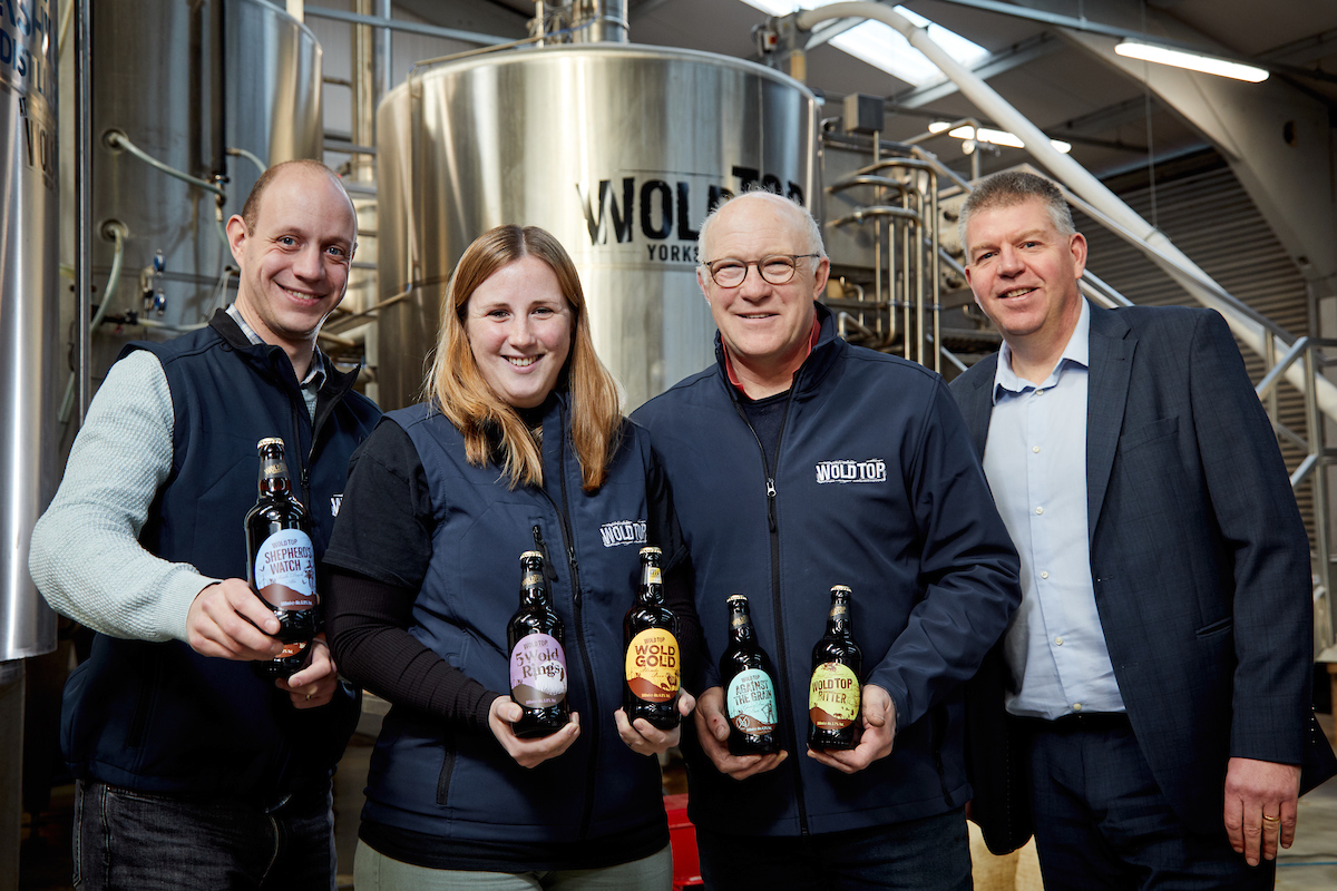 Four people standing in a brewery. There is one woman in the group. They are all holding a bottle of beer apart from a man wearing a navy jacket and blue shirt to the far right.