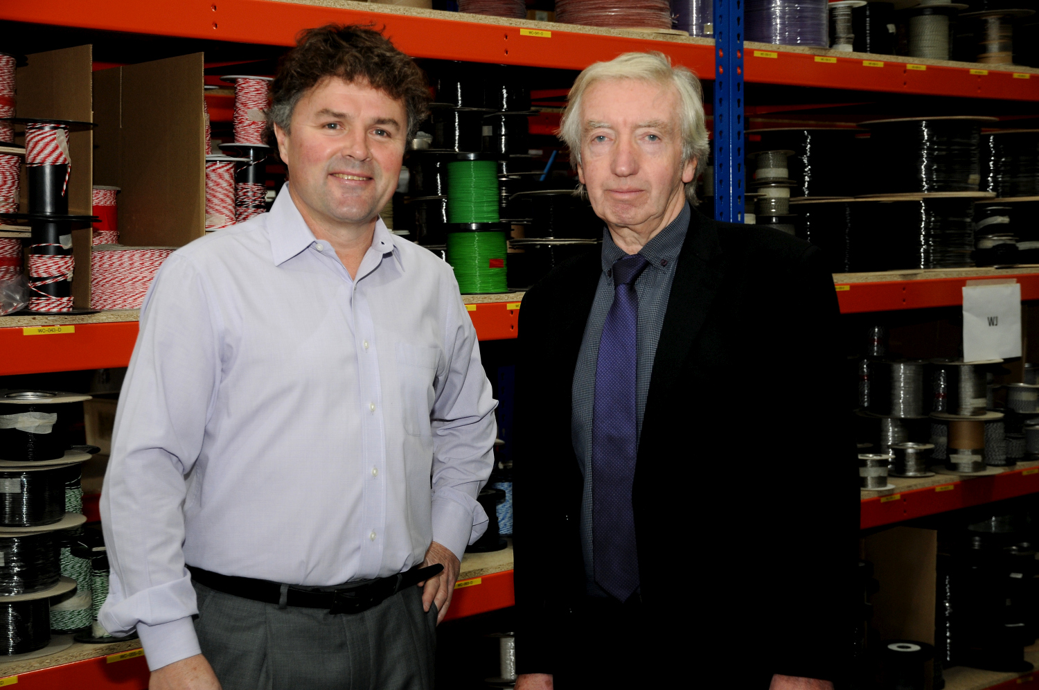 Two men in an industrial unit. Grey haired man to the far left is wearing a black suit, dark grey shirt and navy tie and the other man has dark hair, grey trousers and light blue shirt.