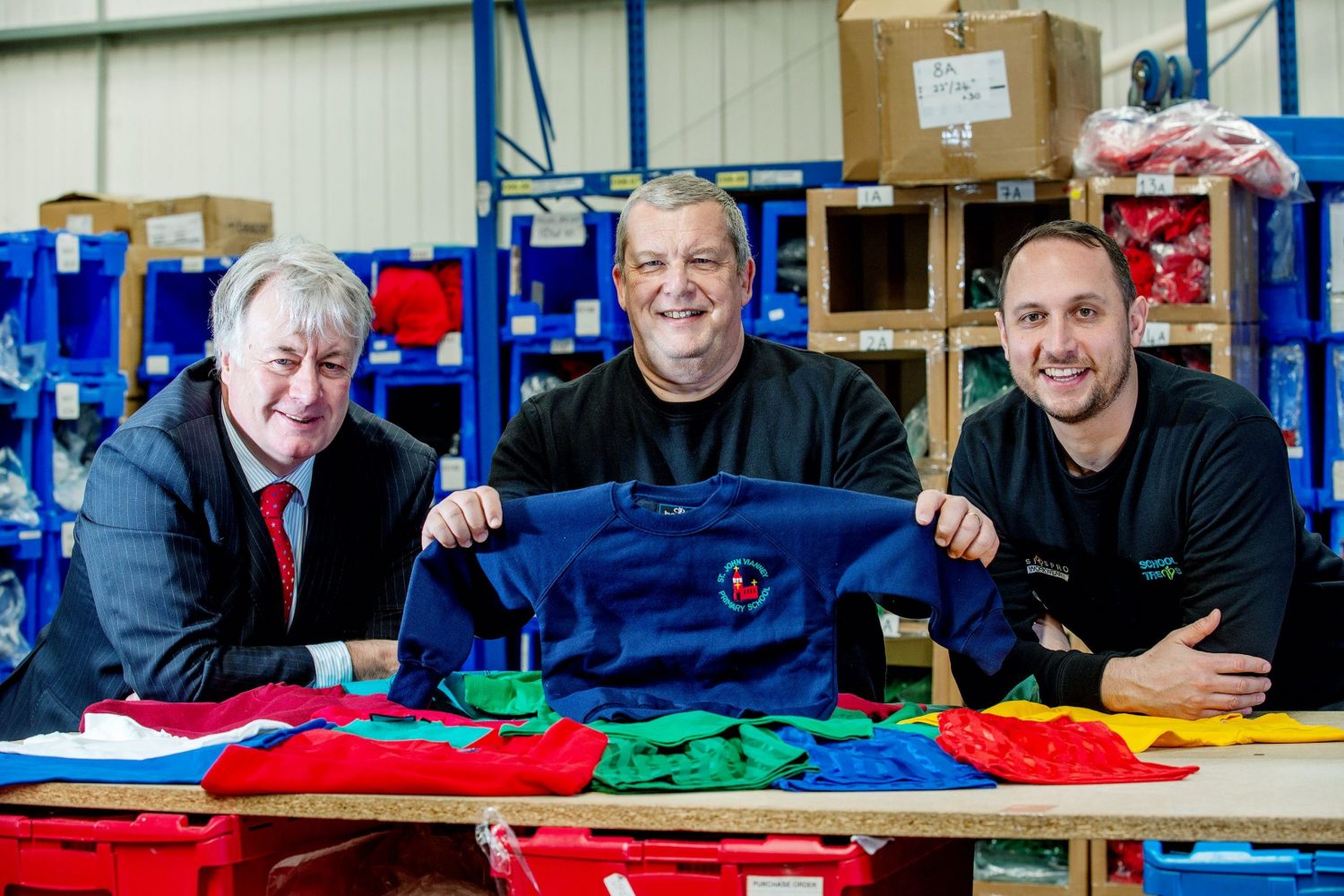 Three men in a clothing factory, man in the middle holding up a blue sweat shirt with a red logo, bearded man to the right hand side wearing a blue sweat shirt and man at the left hand end wearing a blue suit with a red tie has grey hair.