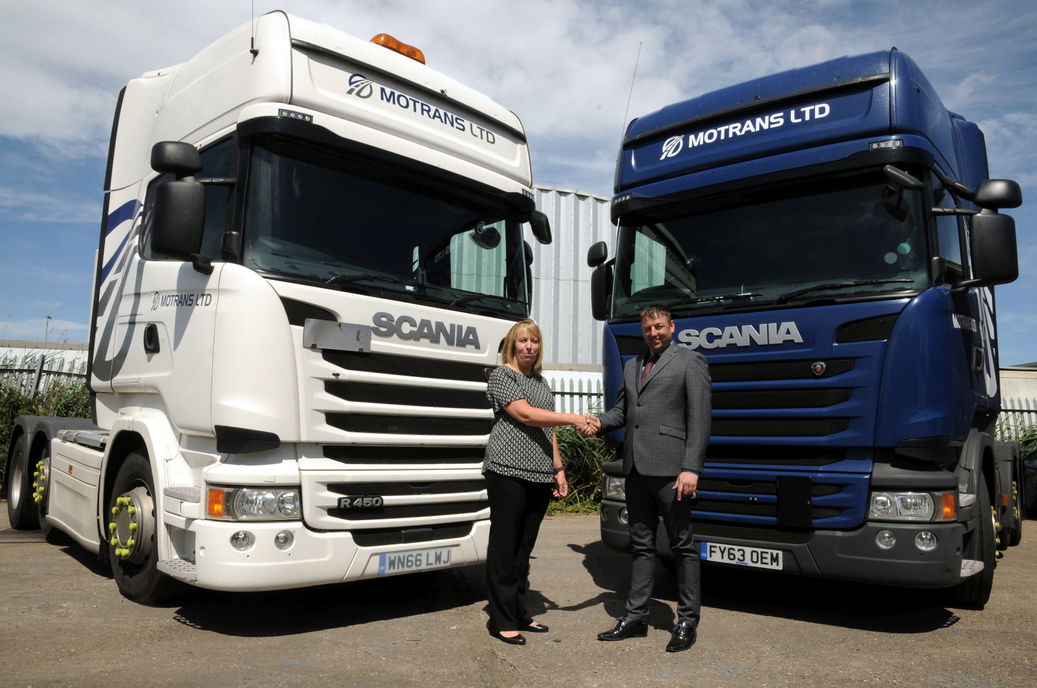 Man and woman shaking hands in front of two Scania trucks. Truck to the far right is dark blue and the other one is white.