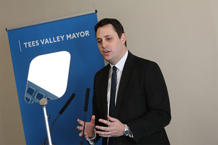 A man in a suit delivering a speech with a Tees Valley Mayor pop up banner in the background