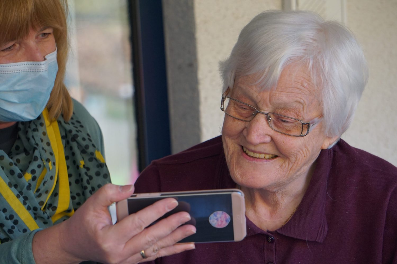 An elderly woman being shown something on a smartphone by a member of care staff wearing a face covering
