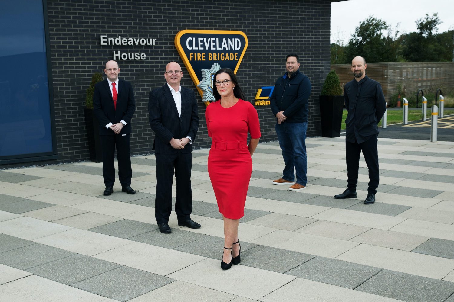 Business woman in a red dress standing in front of four business men. They are outside standing near a brick wall with signage for Cleveland Fire Brigade.